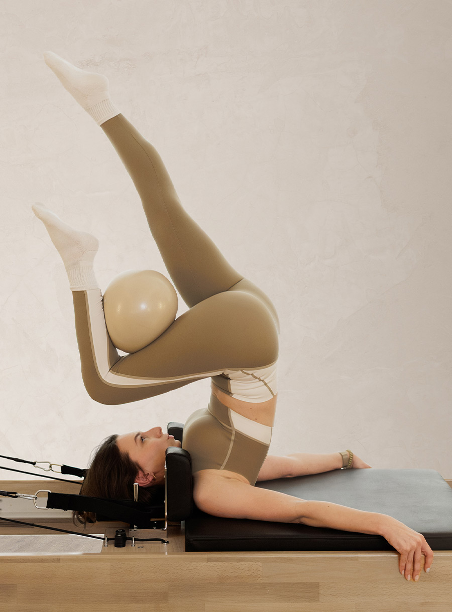 Woman in a tan striped beige pilates outfit holding pilates ball with curled leg while folding lower body over chest